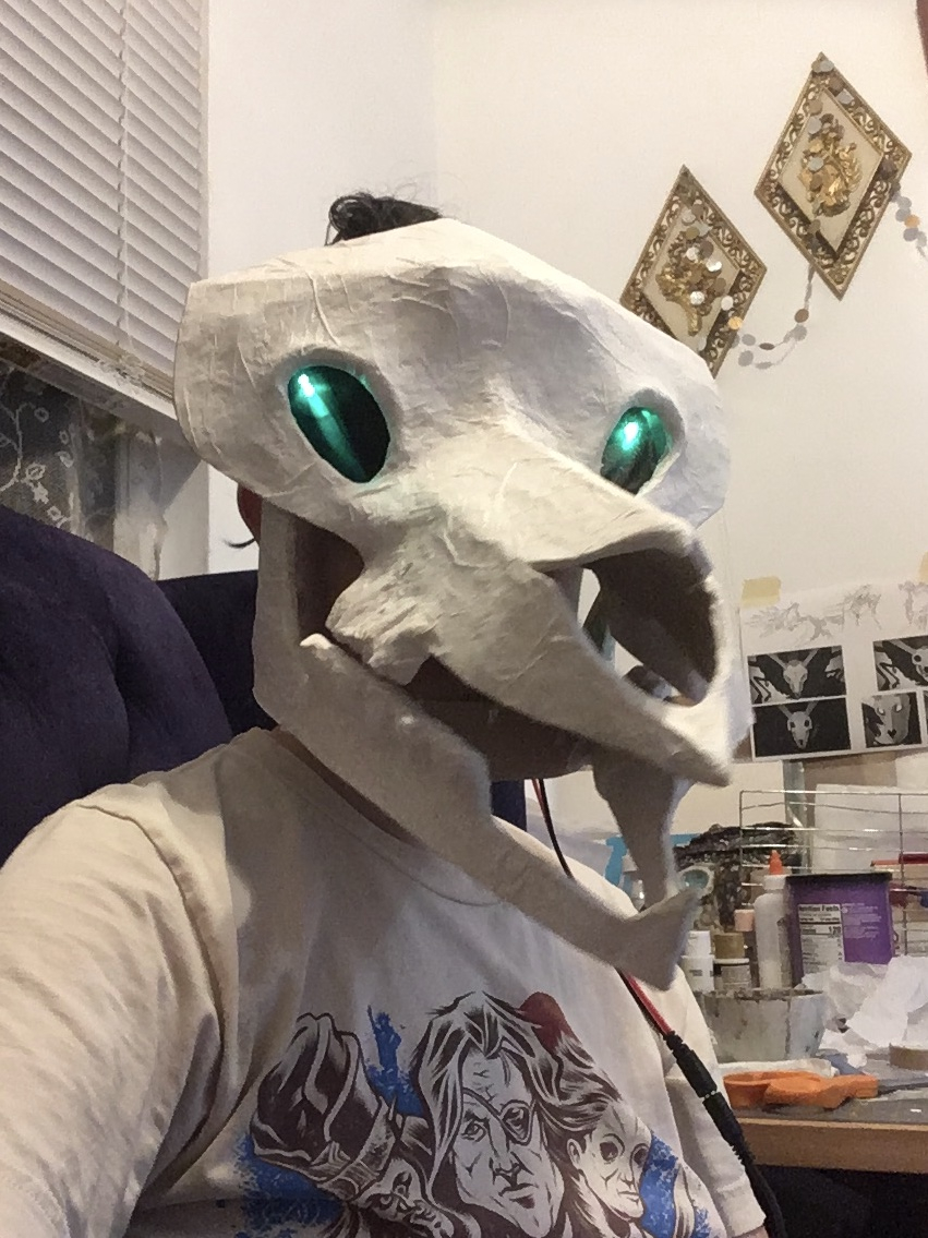 A person sitting on a purple couch, with a messy craft space behind them, wears an oversized, stylised deer skull mask of a rough texture. The eyes are pits of black with bright green lights shining vertically within.