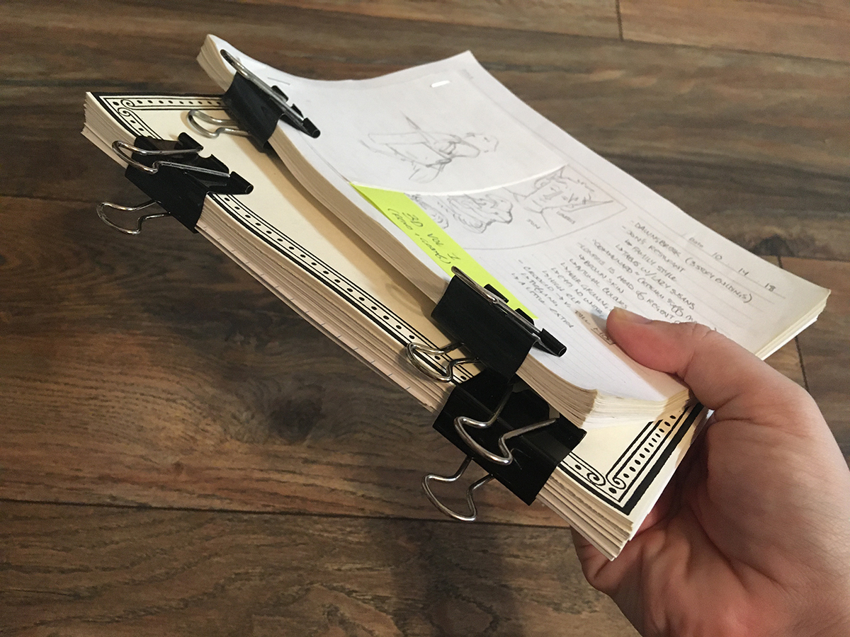 A hand holds two text blocks of sketchbook pages, the sides facing the camera have been squared up and have beefy binder clips holding them together.