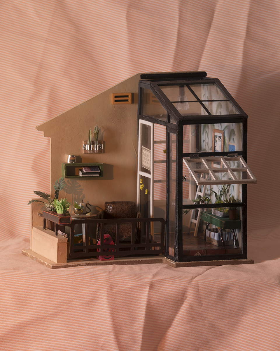 A wider view of the partial dollhouse previously described, photographed on wrinkled orange and white striped fabric and angled to show the attributes from earlier detail images. The light evokes late afternoon golden hour in the southern US, with shadows thrown by the plants on the patio and most of the atrium in warm shadow