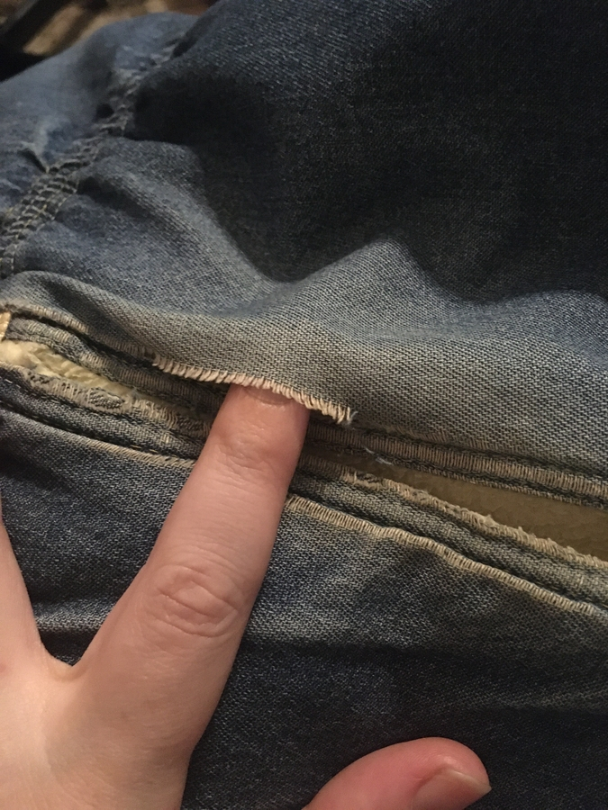 A welt pocket on a lightweight denim coat. Its been worn through where hands brush over the seam. A hand is poking a finger into said hole.