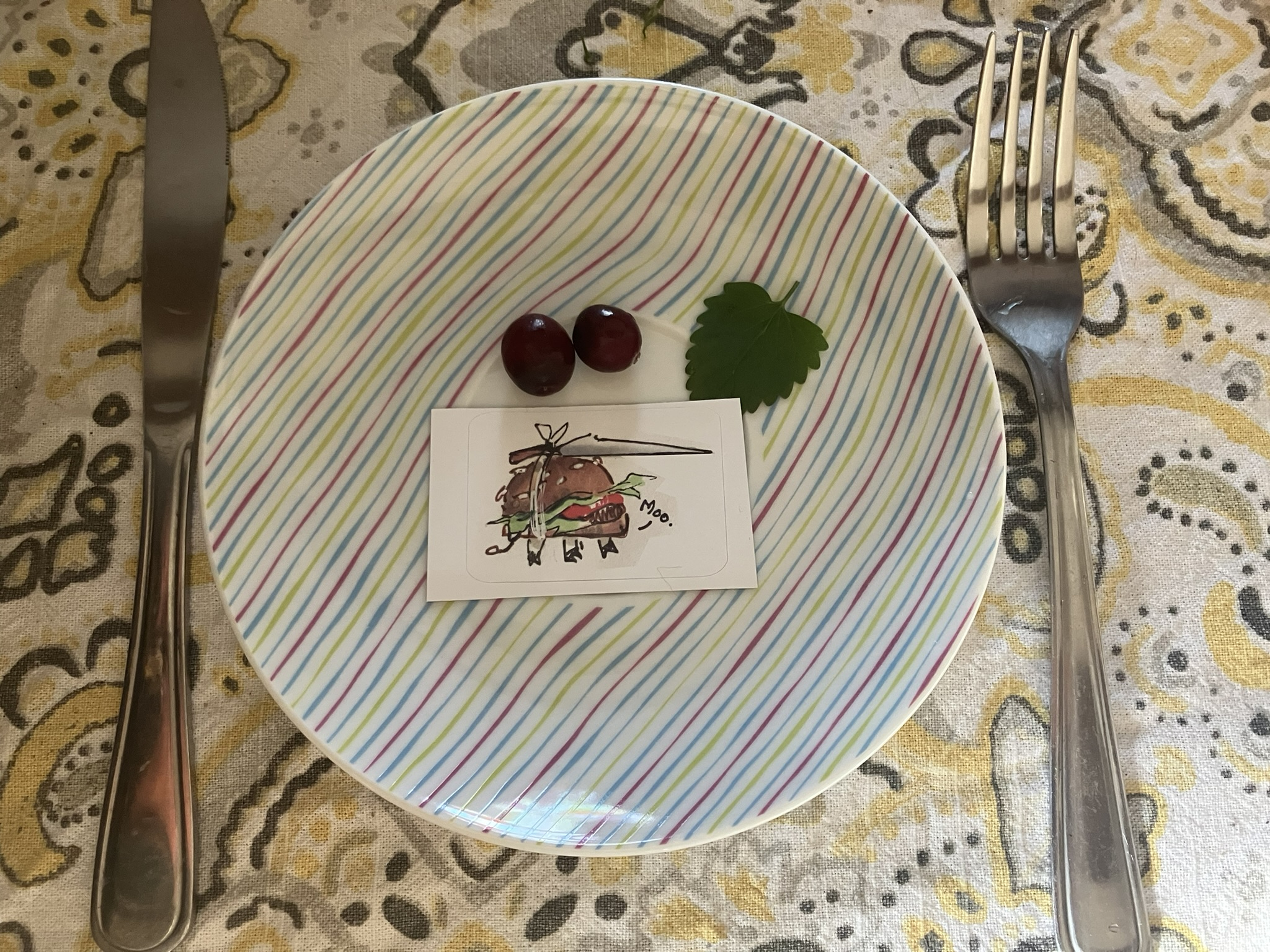 a 'product photo' of the moo burger sticker. it's a hamburger with a knife tied to the top and little cow legs going 'moo' and i've put it on a tiny plate with a garnish (2 cranberries and a catnip leaf) with knife and fork